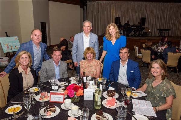 A group of eight well-dressed adults pose for a photo at a formal dinner event. Three individuals are standing behind a round table while five others are seated at the table. The table is set with glasses, plates of cake, salads, rolls, and bottles of wine and water. The background shows a large roo